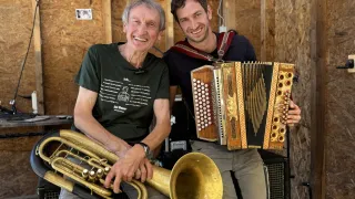 Zwei Männer in einem Studio, einer hält eine Tuba und der andere ein Akkordeon, beide lächeln und posieren für ein Foto.