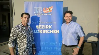 Zwei Männer stehen vor einem blauen Banner mit weißen Text. Einer trägt ein weiß-schwarzes Shirt, der andere ein hellblaues Hemd und khakifarbene Hose. Beide lächeln.
