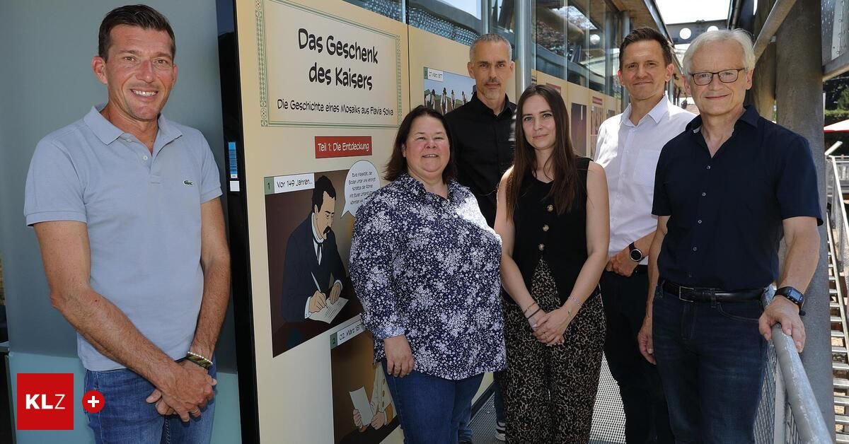Eine Gruppe von Personen steht neben einer Wand mit einem deutschen Schild mit dem Titel 'Das Geschenk des Kaisers.' Sie lächeln für ein Foto. Die Frau auf der linken Seite trägt ein Blumenhemd, während der Mann neben ihr ein Poloshirt trägt. Die Frau in der Mitte trägt ein ärmelloses Top. Die Frau auf der rechten Seite trägt ein schwarzes Kleid.