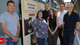 Eine Gruppe von Personen steht neben einer Wand mit einem deutschen Schild mit dem Titel 'Das Geschenk des Kaisers.' Sie lächeln für ein Foto. Die Frau auf der linken Seite trägt ein Blumenhemd, während der Mann neben ihr ein Poloshirt trägt. Die Frau in der Mitte trägt ein ärmelloses Top. Die Frau auf der rechten Seite trägt ein schwarzes Kleid.