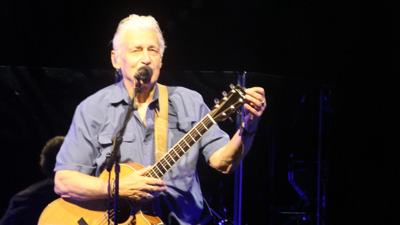 Ein älterer Mann auf der Bühne mit einer Gitarre spielt und singt in ein Mikrofon. Er trägt ein blaues Hemd und hat einen Ring am Finger.