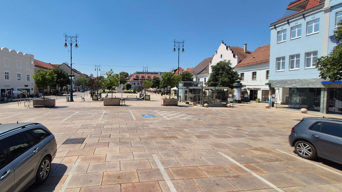 Ein leerer Stadtplatz mit gepflastertem Boden, mehreren Bäumen, Straßenlaternen und Gebäuden im Hintergrund. Ein paar Menschen sind in der Ferne zu sehen.