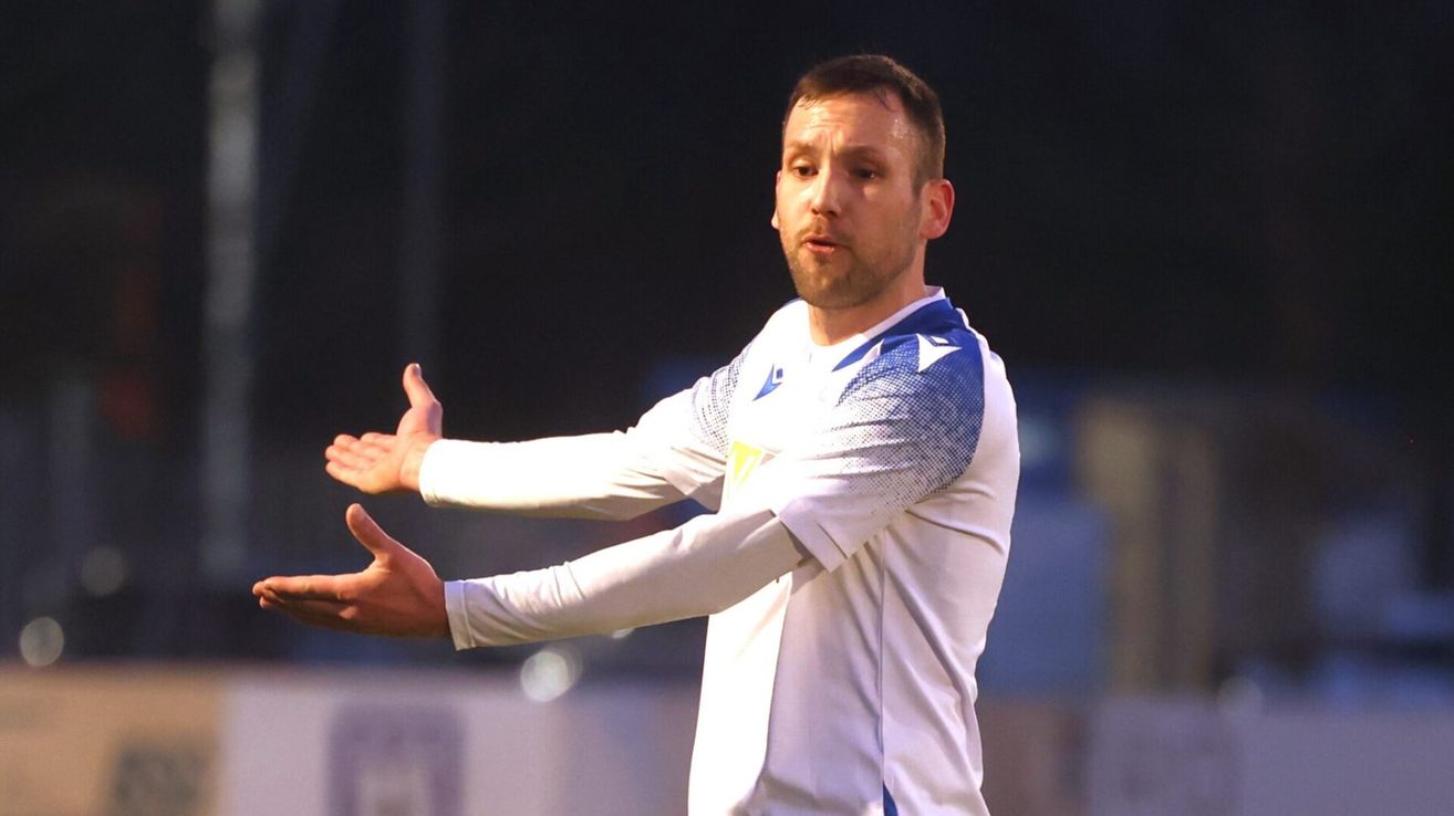 Ein Mann in einem weißen und blauen Trikot gestikuliert mit beiden Händen auf einem Fußballfeld.
