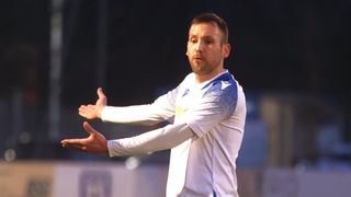 Ein Mann in einem weißen und blauen Trikot gestikuliert mit beiden Händen auf einem Fußballfeld.