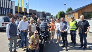 Eine Gruppe von Menschen posiert für ein Foto neben einem Motorrad, mit einem Banner und Gebäuden im Hintergrund.