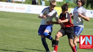 Zwei junge Fußballspieler in blauen und roten Trikots kämpfen auf einem Feld mit grüner Rasenfläche um den Ball.