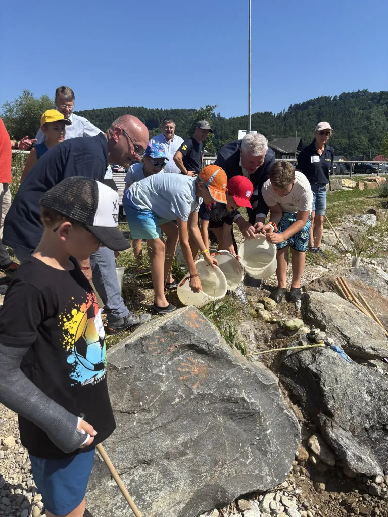 Mehrere Kinder und Erwachsene sind im Freien, wahrscheinlich an einem Fluss, und benutzen Eimer, um im Wasser nach etwas zu suchen. Einige tragen Hüte und Brillen. Bäume und ein Haus sind im Hintergrund.