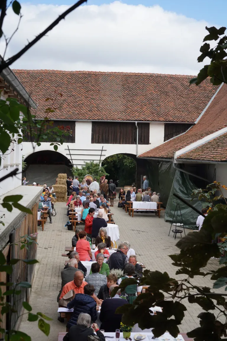 A group of people are gathered for a meal outdoors, under a building with a red roof. Many are seated at tables with white cloths. The area is surrounded by greenery.