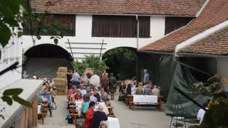 Eine Gruppe von Menschen ist zu einer Mahlzeit im Freien versammelt, unter einem Gebäude mit rotem Dach. Viele sitzen an Tischen mit weißen Tüchern. Der Bereich ist von viel Grün umgeben.