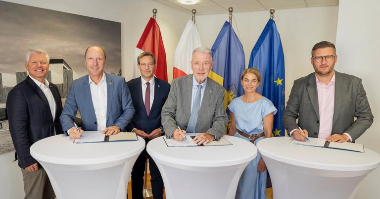 Five individuals stand around a table, signing documents. They are surrounded by flags of various nations, possibly during a formal signing ceremony.