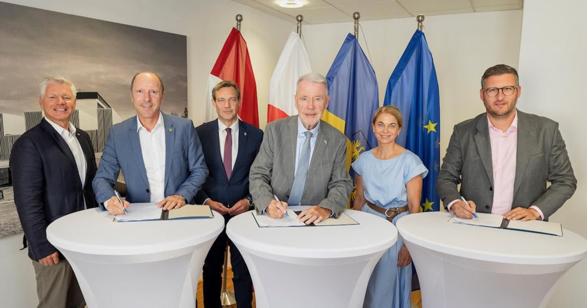 Fünf Personen stehen um einen Tisch und unterzeichnen Dokumente. Sie sind von Flaggen verschiedener Nationen umgeben, möglicherweise bei einer formellen Unterzeichnungszeremonie.