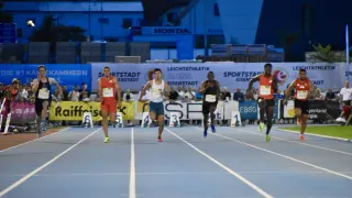 Athleten treten auf einer Laufbahn mit weißen Bahnlinien an. Die Läufer tragen nummerierte Startnummern und treten unter einem Werbebanner an. Zuschauer sehen von den Tribünen zu.