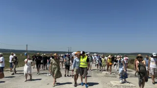 Eine Gruppe von Menschen, einige mit Hüten und Taschen, laufen auf einem Schotterweg unter einem klaren Himmel. Der Mann in der Mitte trägt eine Warnweste.