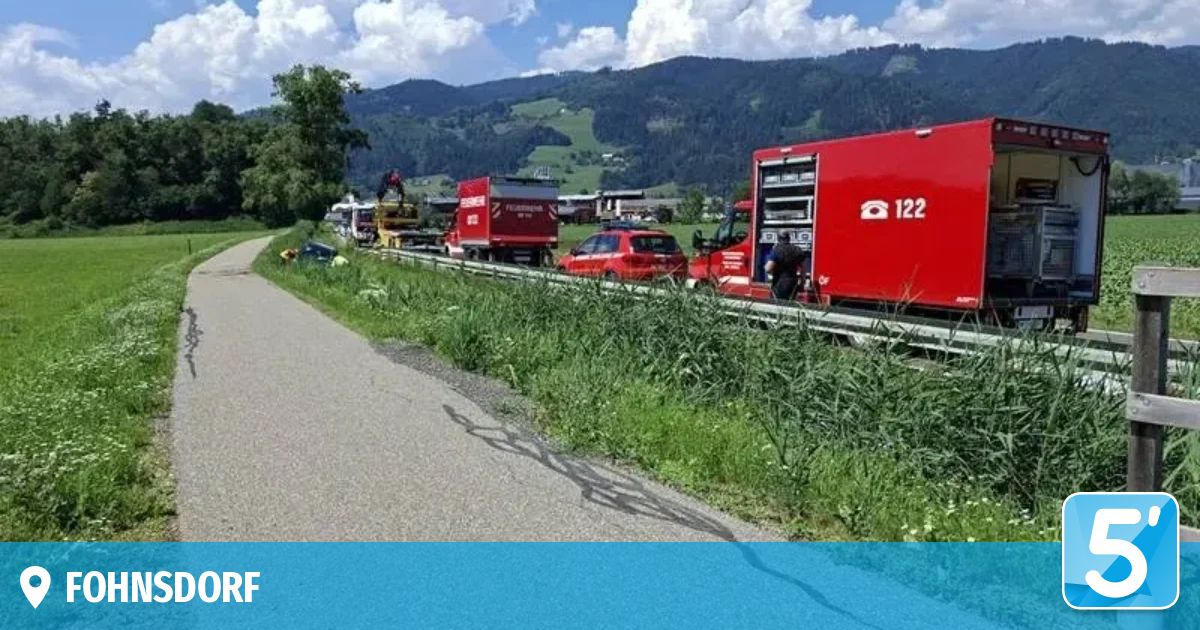 Rettungsfahrzeuge, einschließlich eines roten Feuerwehrwagens, sind am Straßenrand geparkt, mit Bergen im Hintergrund. Arbeiter scheinen einen Unfall zu bearbeiten.