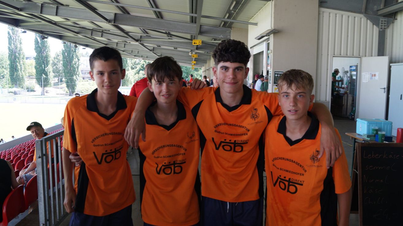 Vier Jungen in orangefarbenen Shirts mit Text darauf, stehen eng beieinander und lächeln. Der Junge auf der rechten Seite hat seine Arme um zwei Jungen gelegt. Hinter ihnen ein verschwommener Hintergrund mit Menschen und einem gelben Schild.