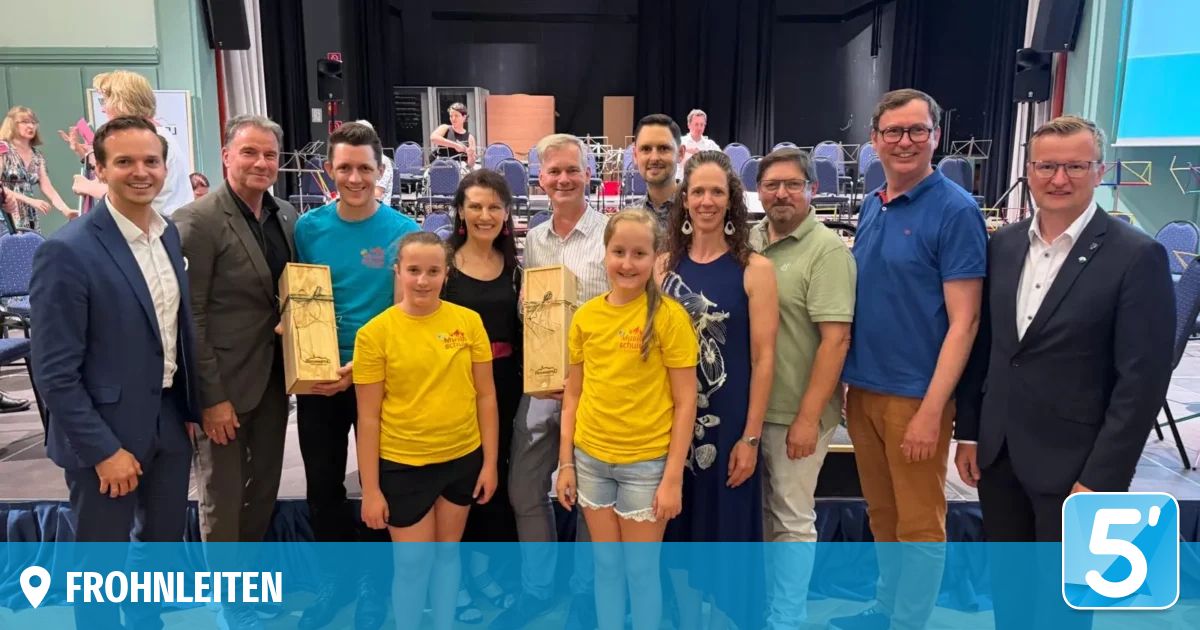 Eine Gruppe von Erwachsenen und Kindern steht auf der Bühne und hält Auszeichnungen. Zwei junge Mädchen in gelben T-Shirts stehen in der Mitte. Dahinter sitzen mehrere Personen auf Stühlen. Ein Wasserzeichen mit der Aufschrift 'TEN' befindet sich links unten.