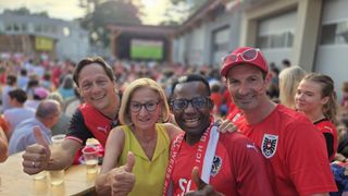 Vier Personen, zwei Männer und zwei Frauen, lächeln und posieren für ein Foto im Freien. Sie tragen alle rote Trikots, wahrscheinlich für eine Sportveranstaltung. Ein Bierglas steht auf einem Tisch im Vordergrund.