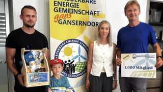 Eine Frau, ein Junge und ein Mann stehen vor einem Schild mit der Aufschrift 'Neuebare Energie Gemeinschaft Gans Ganserndor'. Der Junge hält eine Box und der Mann ein Schild. Die Frau lächelt.