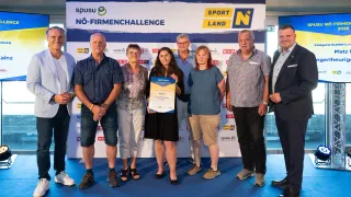 Eine Gruppe von Menschen, darunter zwei Frauen und vier Männer, steht vor einem blauen Teppich und einem Banner. Sie lächeln und posieren für ein Foto. Eine Frau hält eine Urkunde und eine andere Frau einen Ordner.