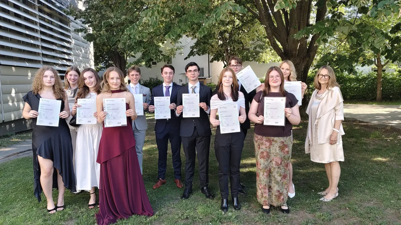Eine Gruppe von Studenten in formeller Kleidung posiert für ein Foto draußen. Sie halten Zertifikate in der Hand und stehen vor einem Gebäude.