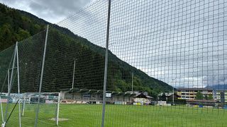 Ein Fußballfeld mit Netzzaun, grünem Rasen und einem Berg im Hintergrund unter einem bewölkten Himmel.