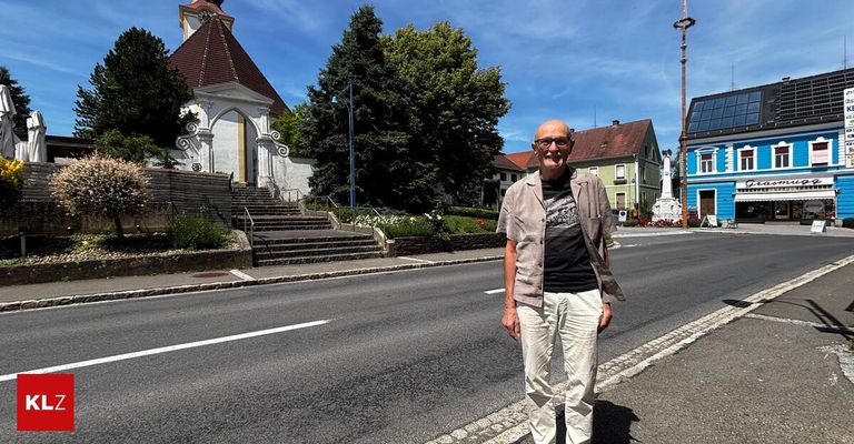 Ein Mann steht am Straßenrand, trägt eine Brille und lächelt, mit einer Kirche und Häusern hinter ihm.