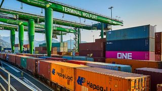 Ein Güterzug fährt in ein Terminal ein, mit gestapelten Containern, gekennzeichnet mit 'Hapag-Lloyd' und 'ONE'. Über Kopfkräne sind bereit, Container zu heben. Das Schild lautet 'Terminal Wolfurt'.
