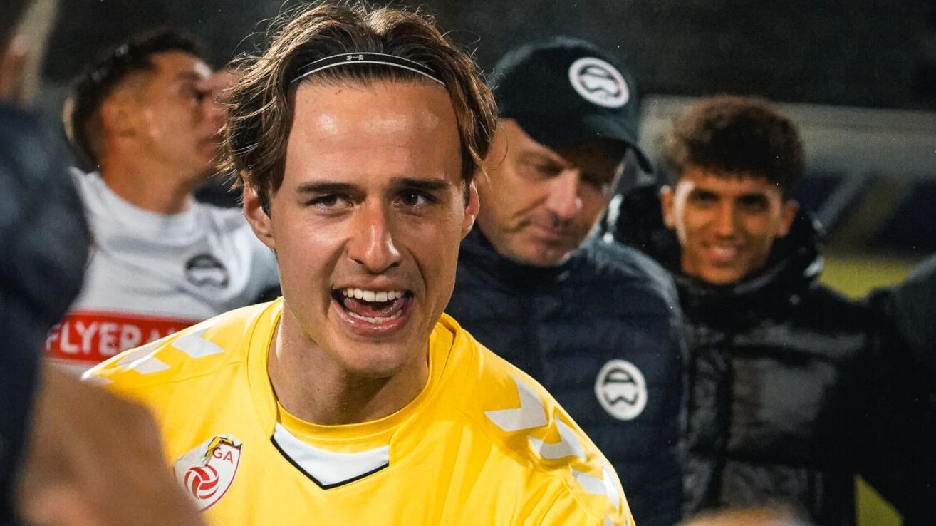 Ein Fußballspieler in einem gelben Trikot mit GA-Logo, schreit und steht mit verschwommenen Teamkollegen im Hintergrund.