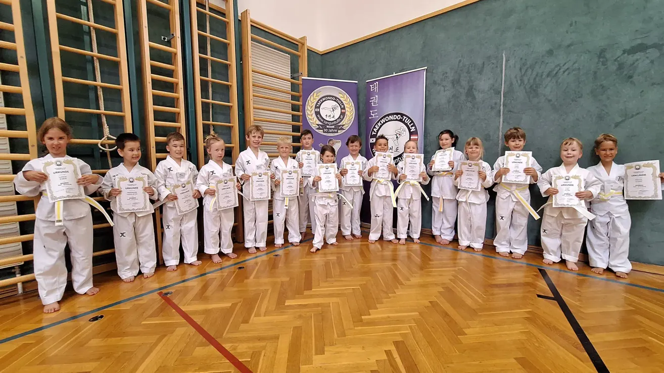Eine Gruppe junger Kampfsportler in weißen Uniformen und gelben Gürteln, die Zertifikate halten, posiert für ein Foto in einer Sporthalle.
