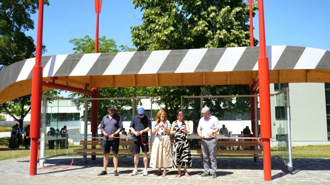 Fünf Personen in Freizeitkleidung halten ein Band und stehen unter einer Holzkonstruktion, möglicherweise für eine Bandzeremonie. Der Hintergrund zeigt Bäume, ein Gebäude und eine Bank.