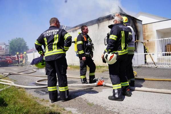 Feuerwehrleute stehen vor einem brennenden Gebäude mit sichtbarem Rauch und Feuer. Die Feuerwehrleute tragen Uniformen und Helme, einer hält eine Feuerwehrspritze.