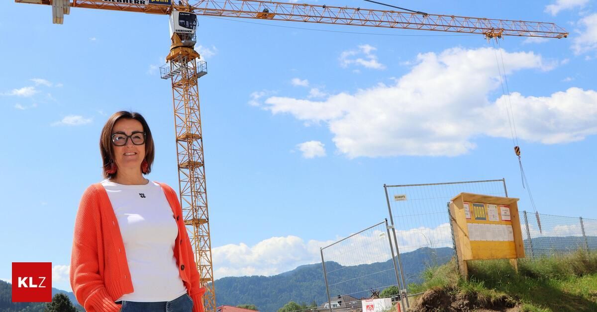 Eine Frau mit Brille und einem orangefarbenen Schal posiert vor einer Baustelle, mit einem hohen Kran und Bergen im Hintergrund.