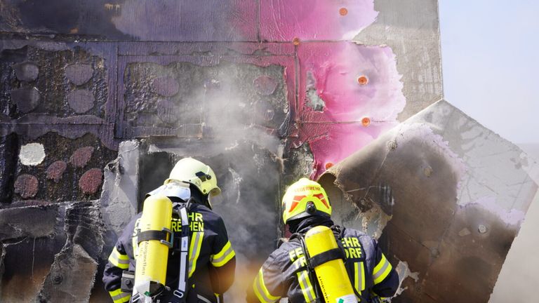 Two firefighters are standing in front of a damaged building, with smoke and pink paint visible on the wall.