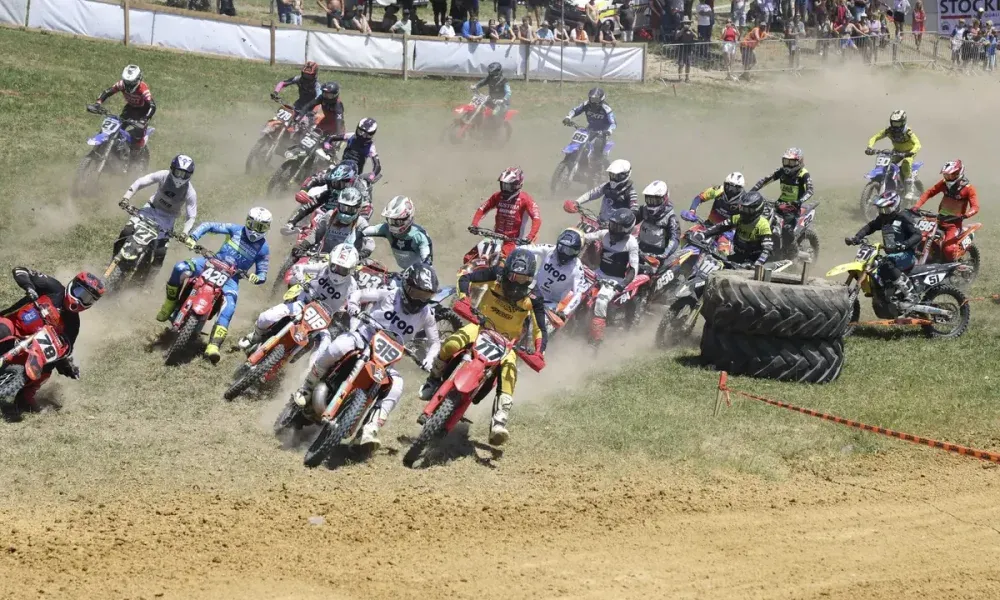 Eine Gruppe von Motorradfahrern in Helmen und Rennanzügen fährt auf einer Dirt-Rennstrecke, während Zuschauer zusehen. Die Fahrer erzeugen Staubwolken und navigieren um Hindernisse. Der Hintergrund zeigt eine verschwommene Menge von Zuschauern.