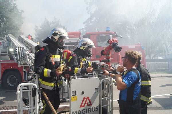 Feuerwehrleute stehen neben einem Feuerwehrwagen mit Rauch im Hintergrund. Sie sind in voller Ausrüstung und halten Ausrüstung. Ein Mann gestikuliert mit der Hand.