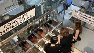 Vier Frauen beobachten einen Schokoladenproduktionsprozess in einer glasverkleideten Fabrik. Ein Schild zeigt Conchierzeitmaschine auf der Glasbarriere. Ein Monitor zeigt Conchieren an.