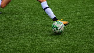Ein Fußballspieler tritt einen Fußball auf einem grünen Kunstrasenplatz. Der Spieler trägt weiße Socken und eine blaue Shorts.
