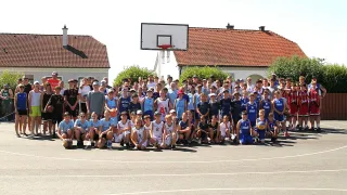 Ein Gruppenfoto einer Jugend-Basketballmannschaft in blauen Trikots, vor einem Basketballfeld mit einem Haus im Hintergrund posierend.