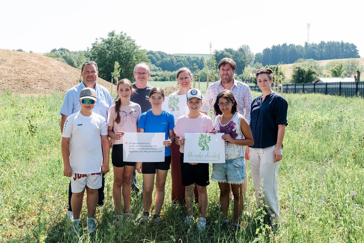 Eine Gruppe von Menschen, einschließlich Kindern, posiert für ein Foto auf einem Feld mit Bäumen. Sie halten Urkunden in der Hand und stehen nebeneinander.