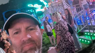 Ein Mann mit Bart und Kappe steht auf einem Jahrmarkt mit einer Menschenmenge um ihn herum. Hinter ihnen befindet sich ein beleuchtetes Fahrgeschäft.