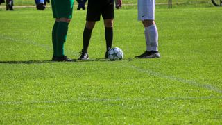 Drei Fußballspieler stehen auf einem Feld, einer hält einen Fußball. Der Spieler in grün und schwarzen Shorts steht links, während der Schiedsrichter in Blau in der Mitte steht.