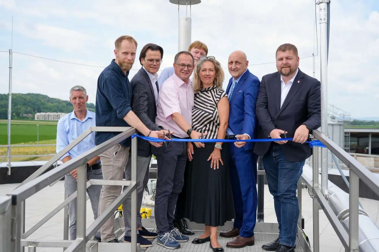 Eine Gruppe von Männern und Frauen steht auf einer Brücke und hält ein blaues Band. Sie lächeln und posieren für ein Foto, möglicherweise für eine Einweihung.
