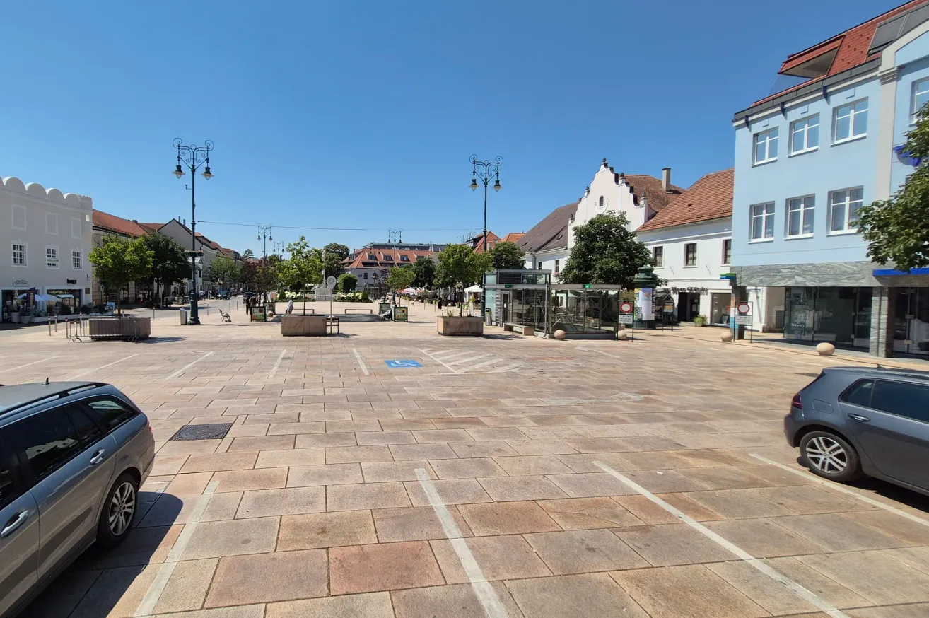 Ein leerer Stadtplatz mit Ziegelpflaster, Straßenlaternen, Bänken und Bäumen. Gebäude mit roten Dächern und blauem Himmel.
