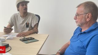Zwei Männer sitzen an einem Tisch, einer mit einer Mütze und einem weißen T-Shirt, der andere in einem blauen Poloshirt und mit Brille, im Gespräch. Papiere und ein Smartphone liegen auf dem Tisch.