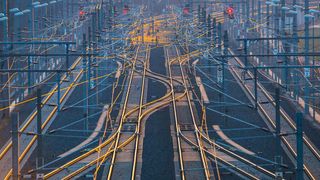 Luftaufnahme eines komplexen Bahnhofs mit mehreren Gleisen und Signalleuchten. Die Gleise sind durch gelbe Linien miteinander verbunden, und die Szene ist in warmes, goldenes Licht getaucht.