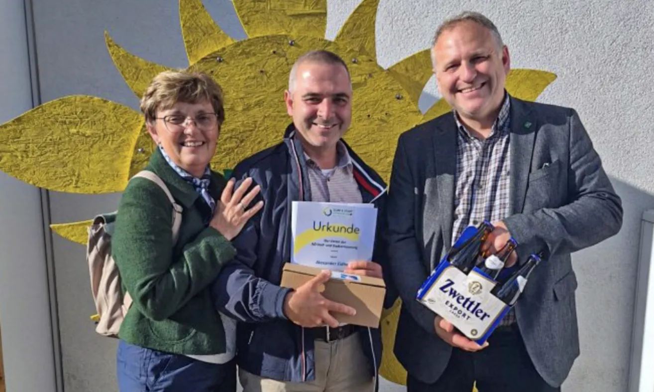 Drei Menschen stehen nebeneinander und lächeln. Ein Mann hält eine Schachtel mit der Aufschrift 'Urkunde' und eine Frau neben ihm hält seine Hand. Ein anderer Mann hält eine Schachtel Zettler Export Bier.