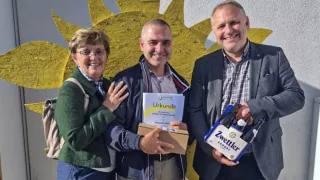Drei Menschen stehen nebeneinander und lächeln. Ein Mann hält eine Schachtel mit der Aufschrift 'Urkunde' und eine Frau neben ihm hält seine Hand. Ein anderer Mann hält eine Schachtel Zettler Export Bier.