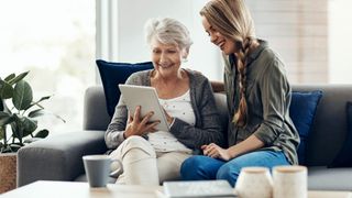 Zwei Frauen sitzen auf einer Couch, die ältere Frau hält ein Tablet und lächelt, während die jüngere sie ansieht. Sie sind von Tassen und Kissen umgeben.