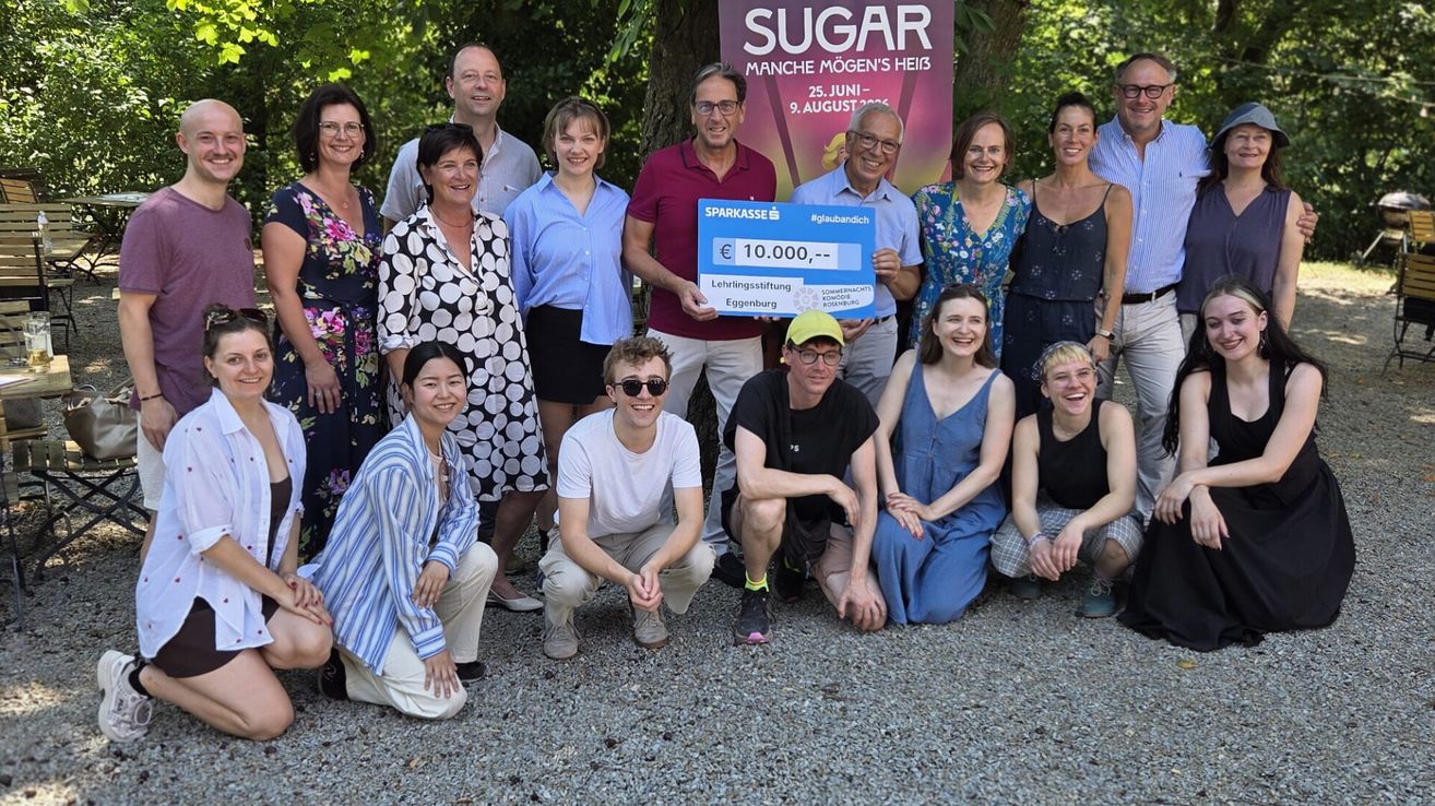 Eine Gruppe von Menschen posiert für ein Foto im Freien. Ein Mann hält einen blauen Scheck mit dem Text 'SUGAR' und '€10.000' und lächelt mit anderen.
