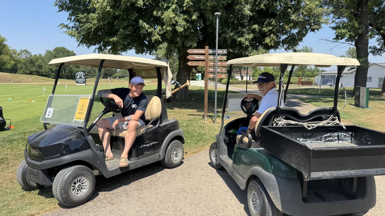Zwei Männer sitzen in Golfcarts, einer lächelt und der andere trägt eine Brille. Ein Baum und Wegweiser sind im Hintergrund.
